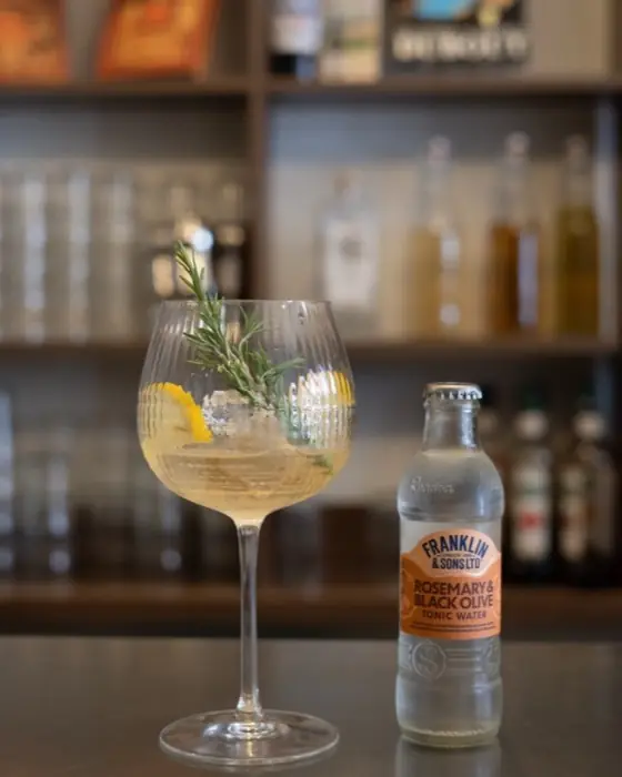 Verre de gin tonic au romarin et au citron, devant les bouteilles du bar.
