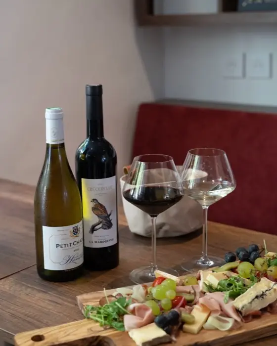 Deux bouteilles, verres de vin rouge et planche de charcuterie, fromages et raisin.